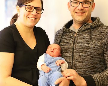 Ein Paar hält ein neugeborenes Baby in den Armen. Die Frau trägt ein schwarzes Oberteil, der Mann ein graues Sweatshirt.