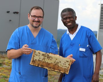 Zwei Männer in blauen Kitteln halten eine Wabe mit Honig in der Hand, während sie neben einem Bienenstock stehen.