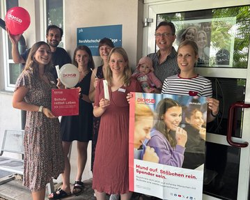 Gruppe von Personen, die für die DKMS werben, halten Plakate und Luftballons. Eine Person hält ein Baby. Hintergrund zeigt ein Gebäude.
