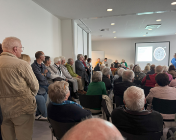 Eine Gruppe älterer Menschen sitzt und steht in einem Raum, während eine Präsentation auf einem Bildschirm gezeigt wird.