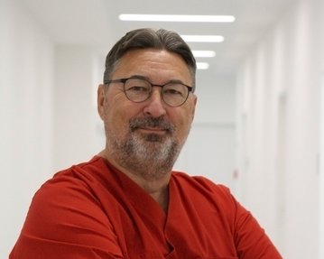 Ein Mann in roter Arztkleidung steht mit verschränkten Armen in einem hellen Flur. Er trägt eine Brille und hat graue Haare.