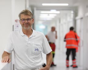 Ein Mann in einem weißen Poloshirt steht in einem modernen Flur, während im Hintergrund Personen in Arbeitskleidung zu sehen sind.