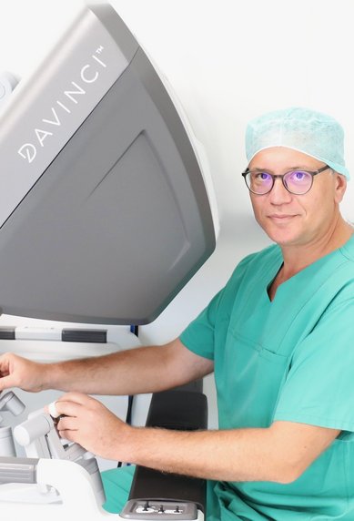Ein Arzt in grüner OP-Kleidung sitzt neben einem Da Vinci Operationsroboter und hält einen Steuerhebel in der Hand.
