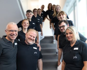Eine Gruppe von 13 Personen in schwarzen Poloshirts steht auf einer Treppe und lächelt in die Kamera.