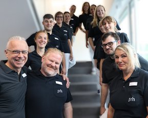 Eine Gruppe von 13 Personen in schwarzen Poloshirts steht auf einer Treppe und lächelt in die Kamera.