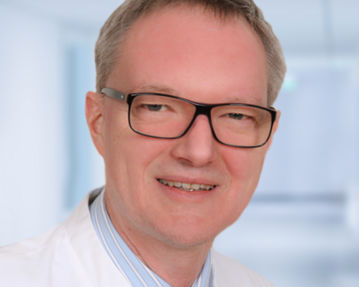 Ein Mann mit Brille und weißem Kittel lächelt in die Kamera. Er trägt ein gestreiftes Hemd und eine rote Krawatte.