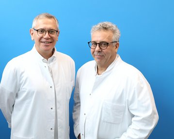 Zwei Männer in weißen Laborkitteln stehen vor einem blauen Hintergrund. Beide tragen Brillen und lächeln leicht.