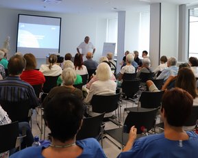 Eine Gruppe älterer Menschen sitzt in einem Konferenzraum und hört einem Vortrag zu, während ein Referent vor einer Präsentation steht.