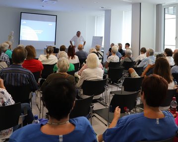 Eine Gruppe älterer Menschen sitzt in einem Konferenzraum und hört einem Vortrag zu, während ein Referent vor einer Präsentation steht.