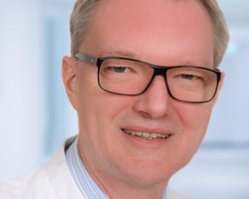 Ein Mann mit Brille trägt einen weißen Kittel und ein gestreiftes Hemd. Er lächelt freundlich in einem hellen, neutralen Hintergrund.