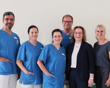 Sechs Personen in medizinischer Kleidung stehen zusammen, lächelnd. Drei tragen blaue Scrubs, während die anderen in formeller Kleidung gekleidet sind.