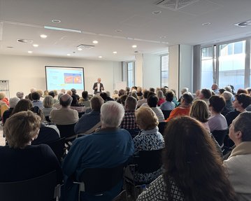 Ein Referent steht vor einem Publikum in einem modernen Raum, während er eine Präsentation auf einem Bildschirm zeigt.