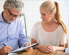 Ein Mann mit Brille schreibt auf einem Notizblock, während eine Frau ihm aufmerksam zuschaut. Beide sitzen an einem Tisch.