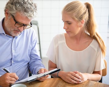 Ein Mann mit Brille schreibt auf einem Notizblock, während eine Frau ihm aufmerksam zuschaut. Beide sitzen an einem Tisch.