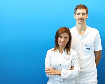 Zwei medizinische Fachkräfte stehen vor einem blauen Hintergrund. Die Frau trägt einen weißen Kittel, der Mann ein weißes Polohemd.