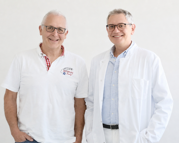 Zwei Männer stehen nebeneinander, einer trägt ein weißes Hemd und der andere einen weißen Laborkittel. Beide lächeln freundlich.