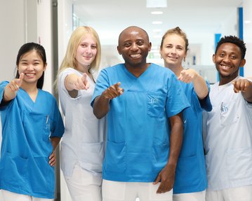 Sechs Personen in blauen Arbeitskleidung stehen in einem Flur und zeigen mit dem Finger auf die Kamera.