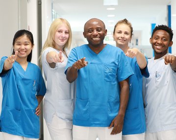 Sechs Personen in blauen Arbeitskleidung stehen in einem Flur und zeigen mit dem Finger auf die Kamera.