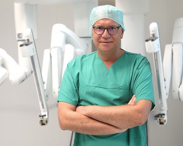 Ein Arzt in grüner OP-Kleidung steht mit verschränkten Armen vor einem chirurgischen Roboter.