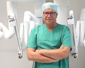 Ein Arzt in grüner OP-Kleidung steht mit verschränkten Armen vor einem chirurgischen Roboter.
