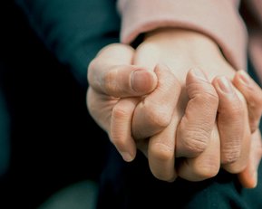 Zwei Hände halten sich fest, eine Hand umschließt die andere. Die Hauttöne sind unterschiedlich, und die Hände sind in einer engen Umarmung positioniert.