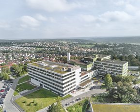 Moderne Gebäude mit begrünten Dächern, umgeben von Parkplätzen und einer städtischen Landschaft. Im Hintergrund sind Wohngebiete und eine hügelige Landschaft sichtbar.