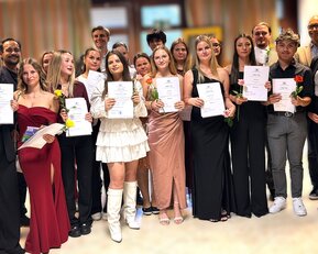 Gruppe von Absolventen hält Urkunden und Blumen, posiert in formeller Kleidung für ein Gruppenfoto bei einer Abschlussfeier.