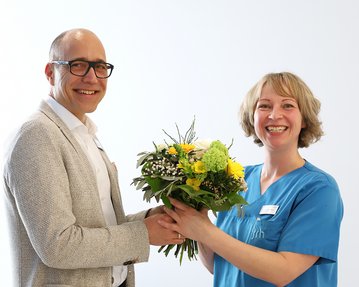 Ein Mann überreicht einer Frau mit blauer Uniform einen Blumenstrauß. Beide lächeln und stehen in einem hellen Raum.