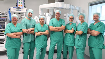Eine Gruppe von sieben medizinischen Fachkräften in grünen OP-Kitteln und Haarnetzen steht vor einem chirurgischen Roboter.