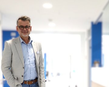 Ein Mann mit Brille und grauem Anzug steht in einem modernen Büro mit hellem Hintergrund.