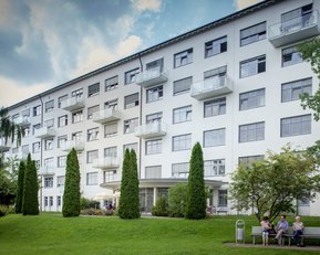 Mehrstöckiges Wohngebäude mit Balkonen, umgeben von Bäumen und Rasen. Im Vordergrund sitzen drei Personen auf einer Bank.