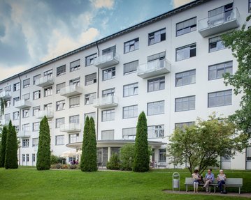 Mehrstöckiges Wohngebäude mit Balkonen, umgeben von Bäumen und Rasen. Im Vordergrund sitzen drei Personen auf einer Bank.