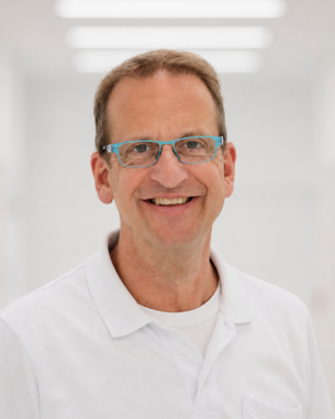 Ein Mann mit Brille und weißem Poloshirt lächelt freundlich in einem hellen, modernen Raum.