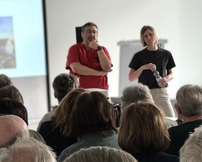 Zwei Personen stehen vor einem Publikum, während eine Präsentation im Hintergrund läuft. Der Mann trägt ein rotes Hemd, die Frau ein schwarzes T-Shirt.