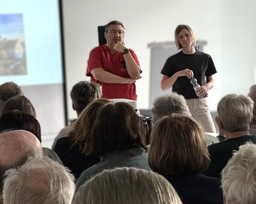 Zwei Personen stehen vor einem Publikum, während eine Präsentation im Hintergrund läuft. Der Mann trägt ein rotes Hemd, die Frau ein schwarzes T-Shirt.