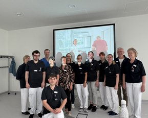 Eine Gruppe von zwölf Personen in Arbeitskleidung steht vor einem Präsentationsbildschirm. Sie lächeln und posieren gemeinsam in einem modernen Raum. Der Bildschirm zeigt Informationen über eine Institution.