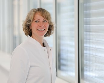 Eine lächelnde Frau in einem weißen Kittel steht neben großen Fenstern. Sie hat lockige Haare und blickt zur Kamera.