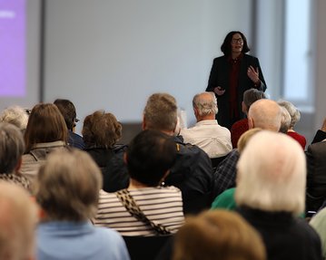 Eine Frau steht auf einer Bühne und spricht vor einem Publikum, das aus älteren Menschen besteht. Im Hintergrund ist eine Präsentation sichtbar.