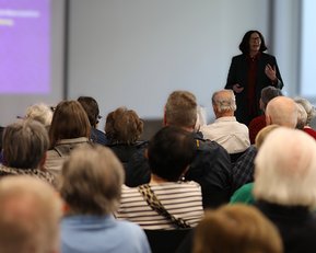 Eine Frau steht auf einer Bühne und spricht vor einem Publikum, das aus älteren Menschen besteht. Im Hintergrund ist eine Präsentation sichtbar.