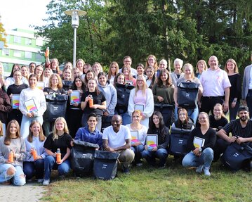 Eine große Gruppe von Menschen steht im Freien, hält Taschen und Urkunden, umgeben von Bäumen und einem Gebäude im Hintergrund.