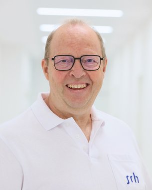 Ein lächelnder Mann mit Brille trägt ein weißes Polohemd mit dem Logo "srh". Der Hintergrund ist hell und unscharf.
