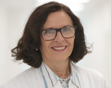 Eine Frau mit lockigem Haar und Brille trägt einen weißen Kittel. Sie lächelt freundlich in einem hellen, modernen Raum.