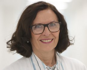 Eine Frau mit lockigem Haar und Brille trägt einen weißen Kittel. Sie lächelt freundlich in einem hellen, modernen Raum.