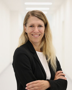 Frau mit langen, blonden Haaren in einem schwarzen Blazer, die lächelnd mit verschränkten Armen in einem hellen Flur steht.
