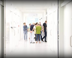 Eine Gruppe von Personen steht in einem hellen Flur mit weißen Wänden und Türen, während sie miteinander sprechen und sich umsehen.