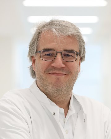 Ein Arzt in einem weißen Kittel lächelt und steht mit verschränkten Armen in einem hellen, modernen Raum.