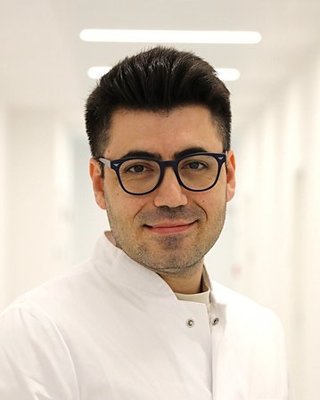 Ein Arzt in einem weißen Kittel steht in einem hellen Flur und lächelt, mit Brille und gepflegtem Aussehen.