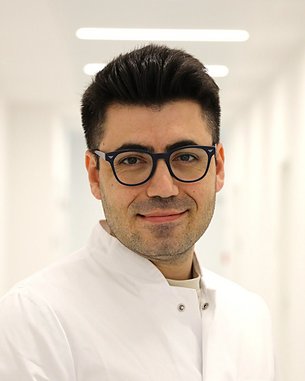 Ein Arzt in einem weißen Kittel steht in einem hellen Flur und lächelt, mit Brille und gepflegtem Aussehen.