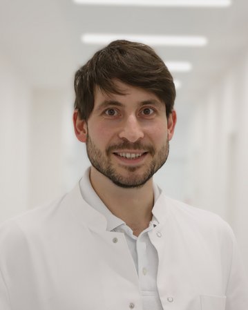 Ein junger Mann mit Bart trägt einen weißen Kittel und lächelt freundlich in einem hellen Flur.