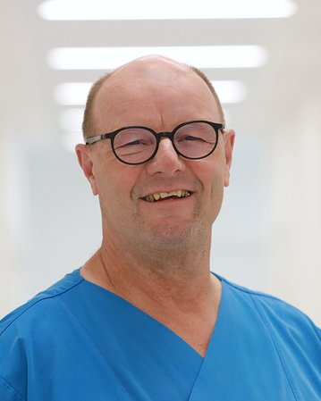 Ein lächelnder Mann in blauer medizinischer Kleidung steht in einem hellen, modernen Flur. Er trägt eine Brille und hat eine Glatze.